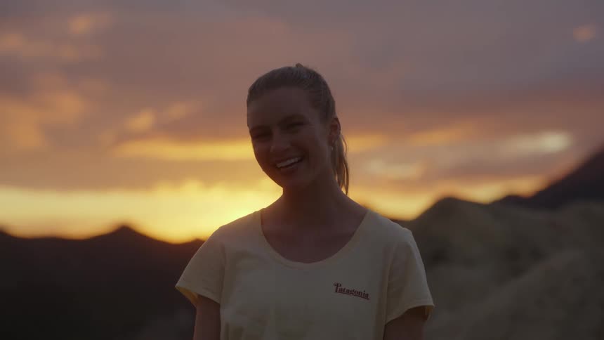 A woman standing atop a mountain at sunset laughs while looking into the camera