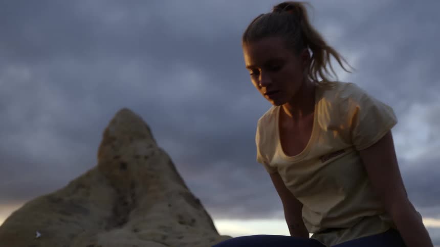 A woman is climbing up the side of a mountain and pulls herself up as she reaches the top