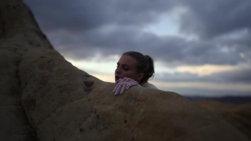 Woman with one hand climbs over the rock at the top of a mountain under a cloudy sky