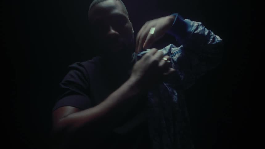 A man is standing in the darkness and he takes off his light blue jacket and turns it inside out and puts it back on