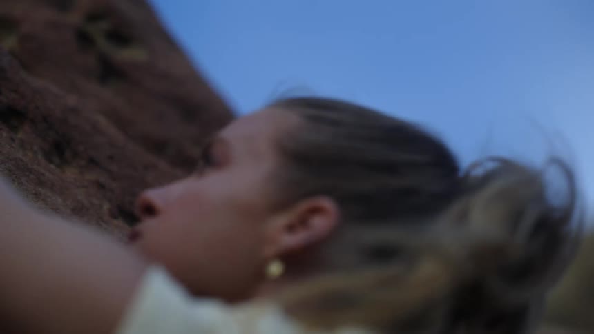 Blonde woman with an earring reaches with her arm to assist her in climbing the rocks on the side of the mountain on a breezy day
