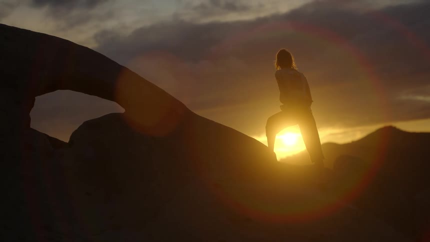 Silhouette of a young woman with a limb difference climbing a rock wall in front of the sun setting over the mountains