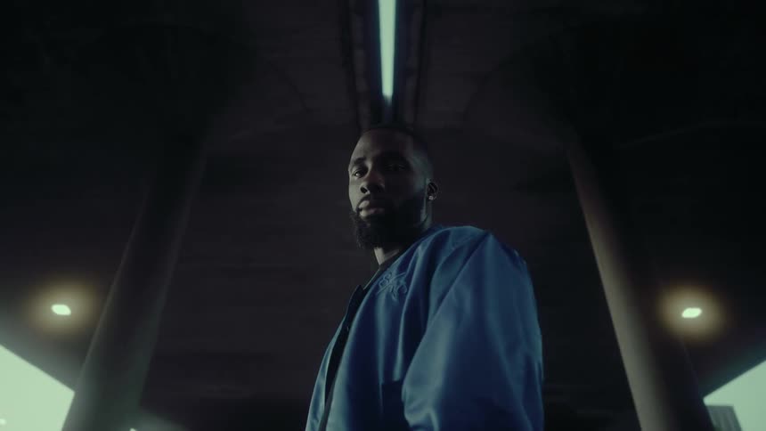 Black man stands on a path under a structure and turns his head to his left as a van travels by on a nearby street
