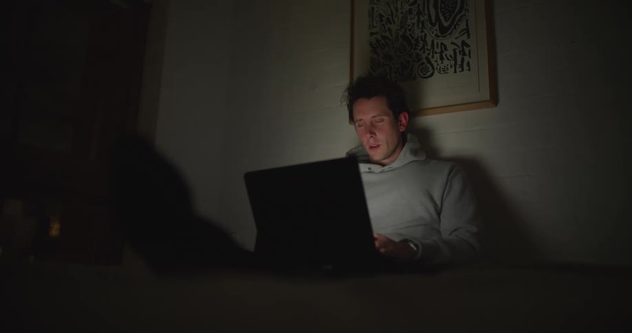 A man is sitting in a dark room leaning against a wall with a laptop and he closes the computer lid