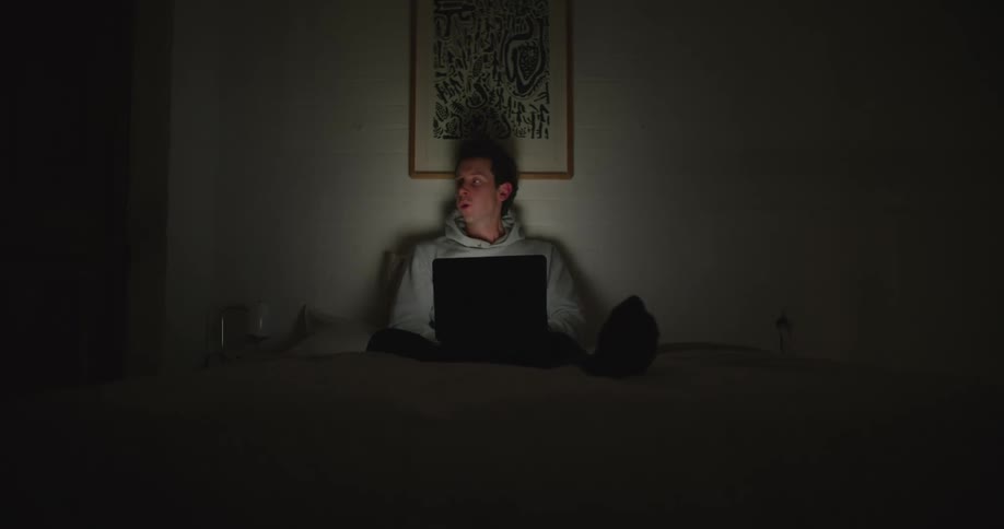 Young tired man sitting in the bed yawns while working at home on a laptop at night