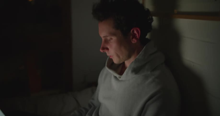 A man typing on a laptop while sitting on his bed at night stops for a moment to run his hand over his face in exhaustion