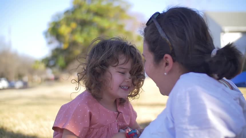 A toddler girl and a woman sit laughing and talking outside in a park