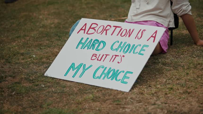 A woman sitting on the grass with a pro choice sign propped against her legs
