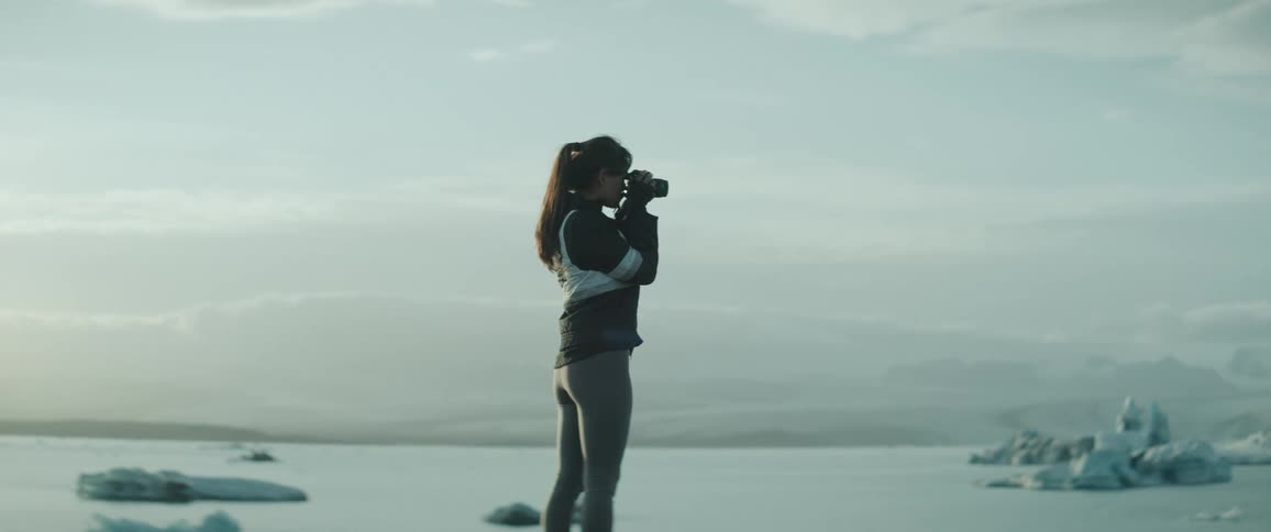 A woman is standing next to an icy lake in the mountains as she takes pictures with her camera