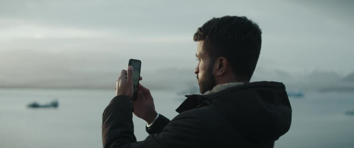A man is holding his cell phone up as he stands on the waterfront of a lake in the mountains