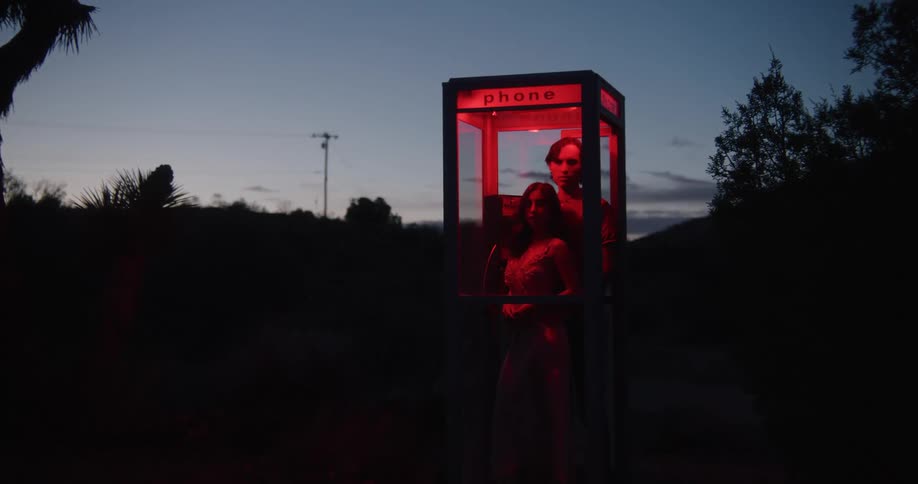 A couple is inside of a phone booth in the forest area at night time