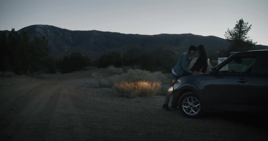 A man picks a woman up and sets on a the hood of a car and they put their heads together and hug