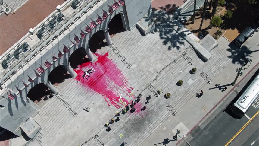 Red paint flows down the stairs in front of a building flying united states of america flags as people hold signs and a bus passes on the street