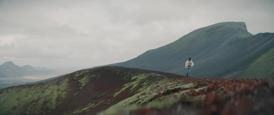 Man with white shirt jeans and boots walks on hill on cloudy day