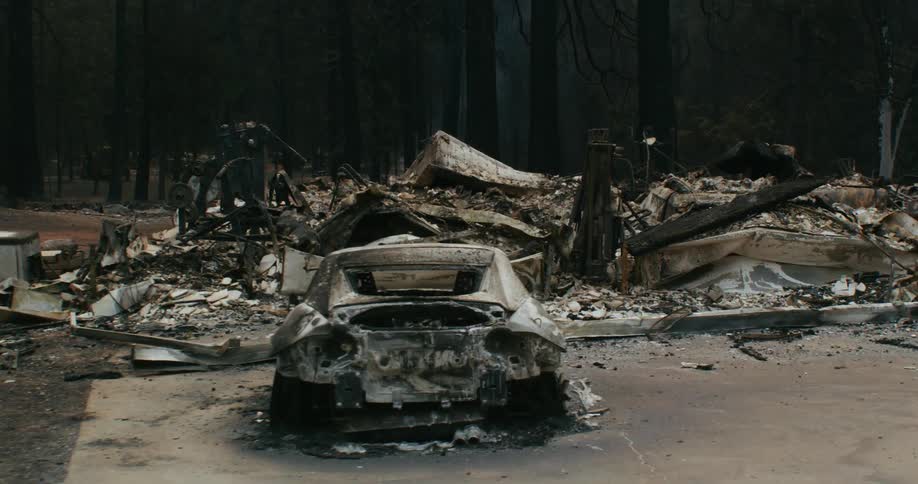A burned out automobile sits in front of a debris from a burned and destroyed building as smoke floats among the trees in the woods