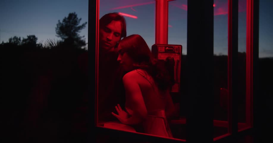 A young woman in a phone booth leans up against the glass where a young man is leaning and they look at one another