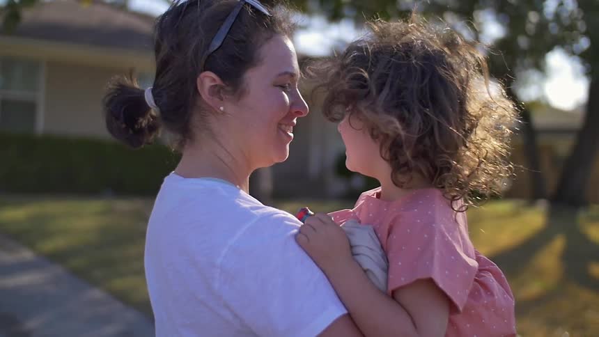 A woman standing out in her front yard holds a toddler who looks upset