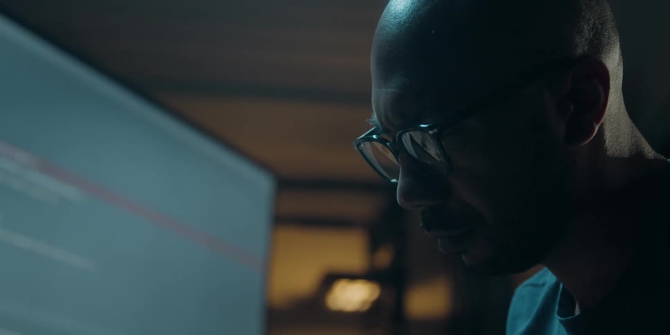A man is sitting at a computer screen indoors and looks down at the desk