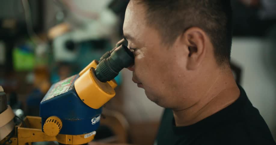 A man is sitting at a table moving something with tweezers and looking at it in a microscope
