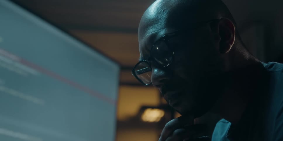 A man sits in front of a computer in a dark office and rubs his neck