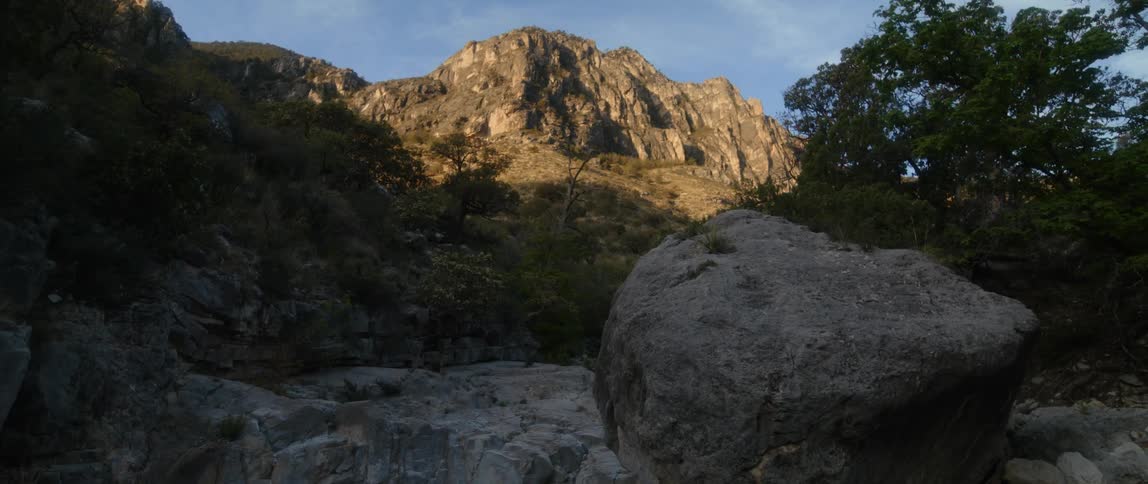 A large boulder and rocks are near trees in the shade as the sun shines on a mountain in the distance