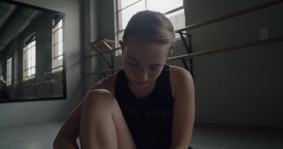 A young ballet dancer sits and ties her shoes in the studio