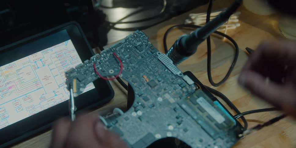 A man is sitting at a table working on an electronic component and he puts the soldering iron into the stand in front of him