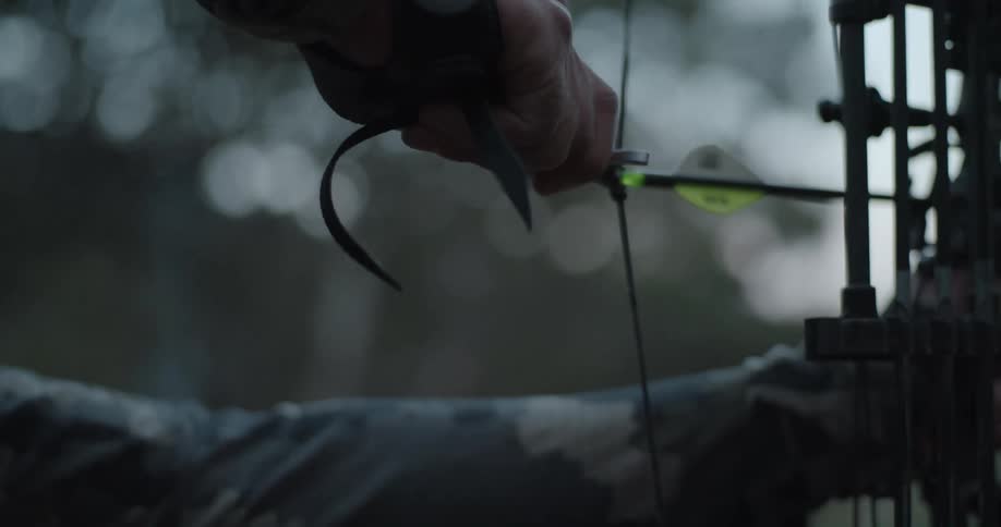 An archer slowly raises a bow while standing outdoors during the day