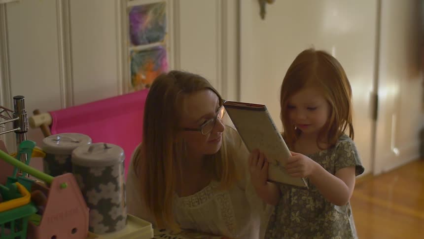 A woman kneels by a girl as she looks at a book