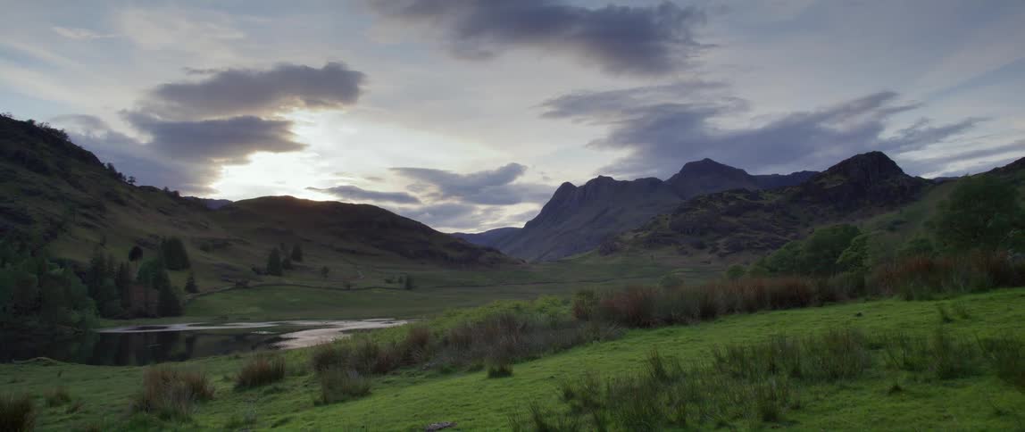 Small body of water in a green field near the mountains under a cloudy blue sky