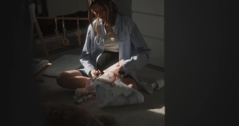 Pregnant woman with brown hair blue and white shirt and black shorts sits on floor cutting cricut stickers with scissors in baby room