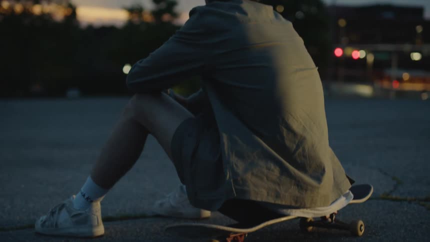 Man with shirt shorts and white sneakers sits on skateboard at dusk