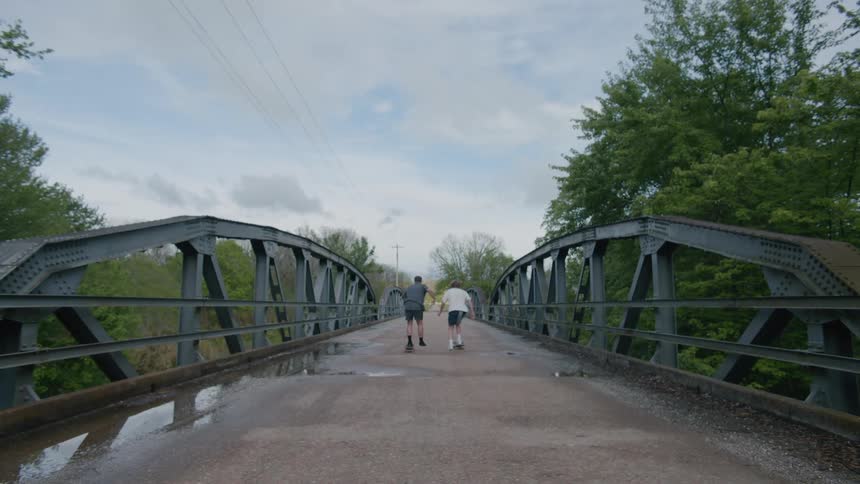 Two teenage boys get a running start as they ride skateboards across a bridge on a forest road