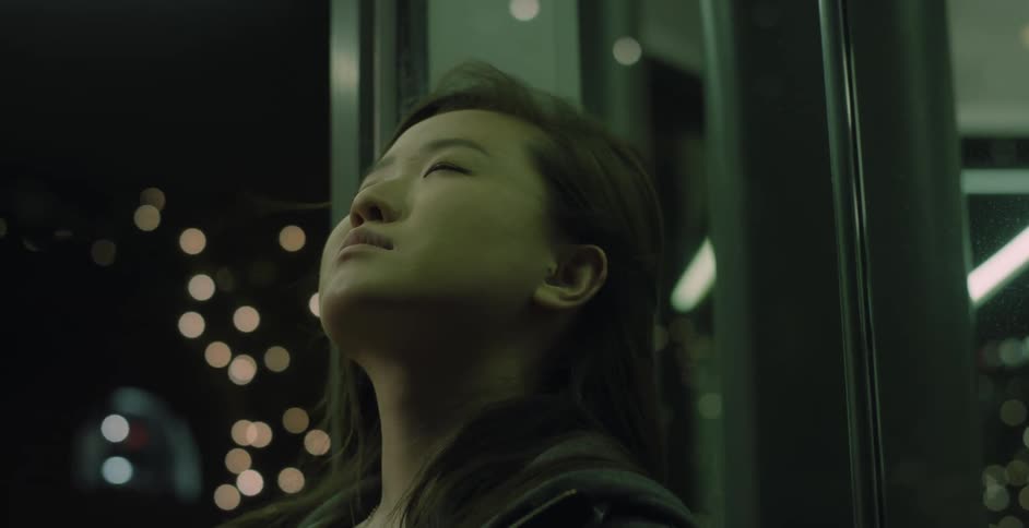 A teenage girl is standing outside on a city street at night leaning against a building and looking up