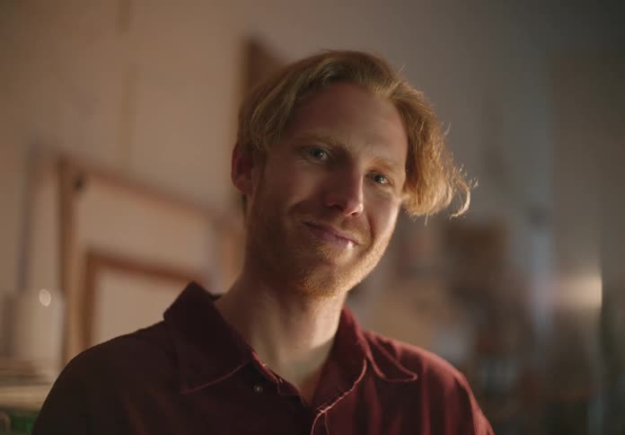 A man is sitting in a room looking down and he looks up and smiles