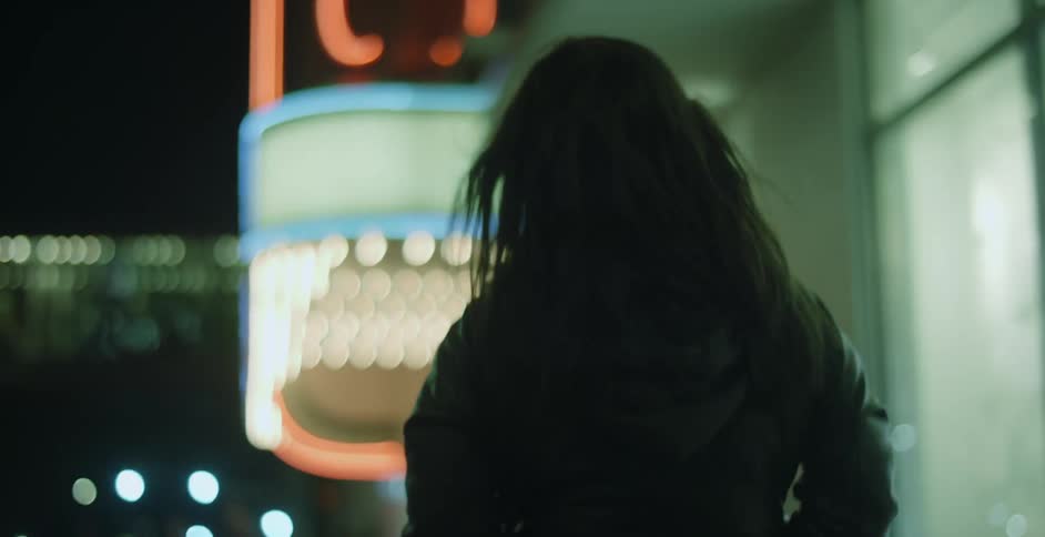A woman is walking on a downtown street at night and her long dark hair blows in the wind