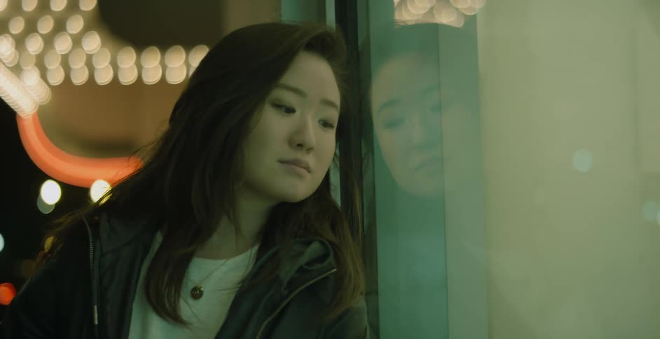 A teenage girl is standing outside on a city street at night leaning against a building and looking sad