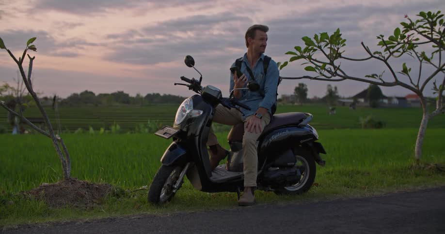 A man is sitting on a moped on the side of a road next to a farm