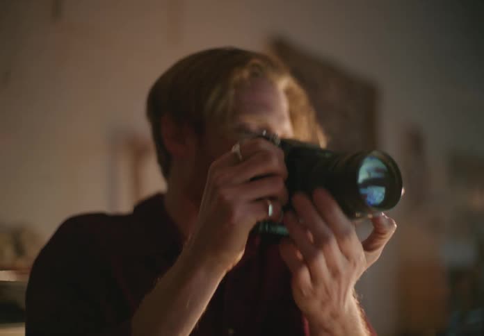 A man at home looks through his camera to take a photo and then examines his camera and adjusts it
