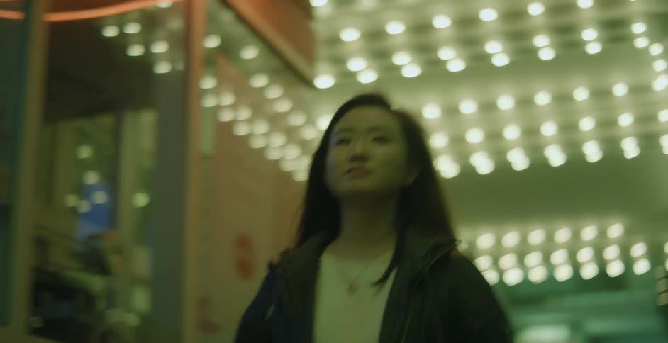 Woman with dark hair necklace and jacket walks down the street next to illuminated building