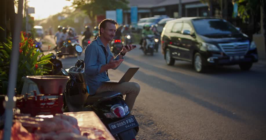 Man sitting on a parked motorcycle a the side of a busy street working on a laptop at evening