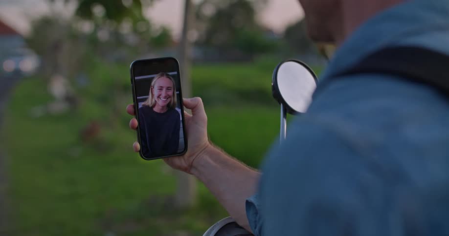 A man is outside sitting on his motorbike and holding his cell phone while he has a video conversation with a woman