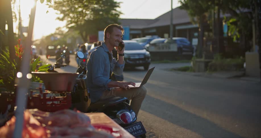 Man sitting on a parked motorcycle a the side of a busy street working on a laptop at evening