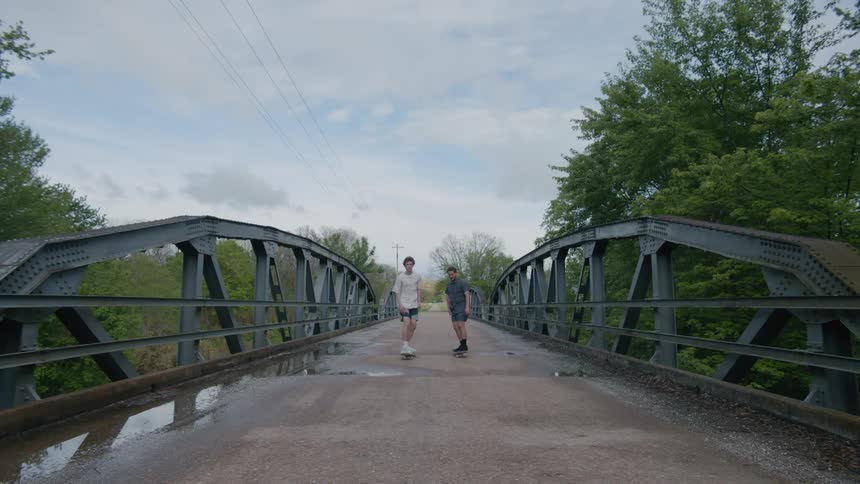 Two men riding skateboards across a pedestrian bridge one attempting and failing a trick
