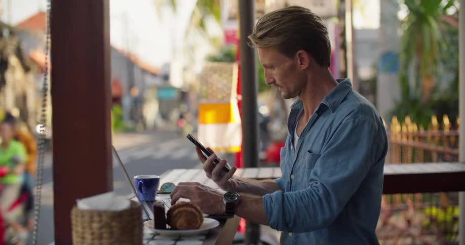 A man is sitting at a streetside cafe typing on a laptop computer and he picks up his cell phone as traffic moves by on the street