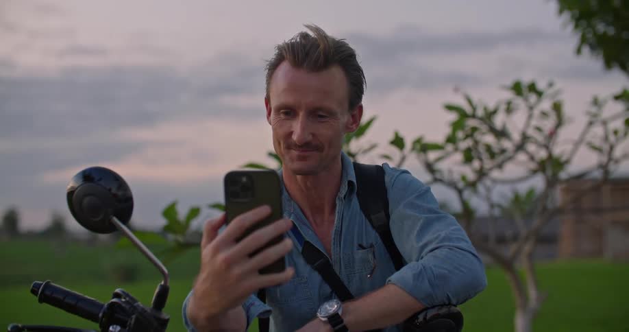 Man with mustache blue shirt and watch sits on motorcycle using smartphone at dusk