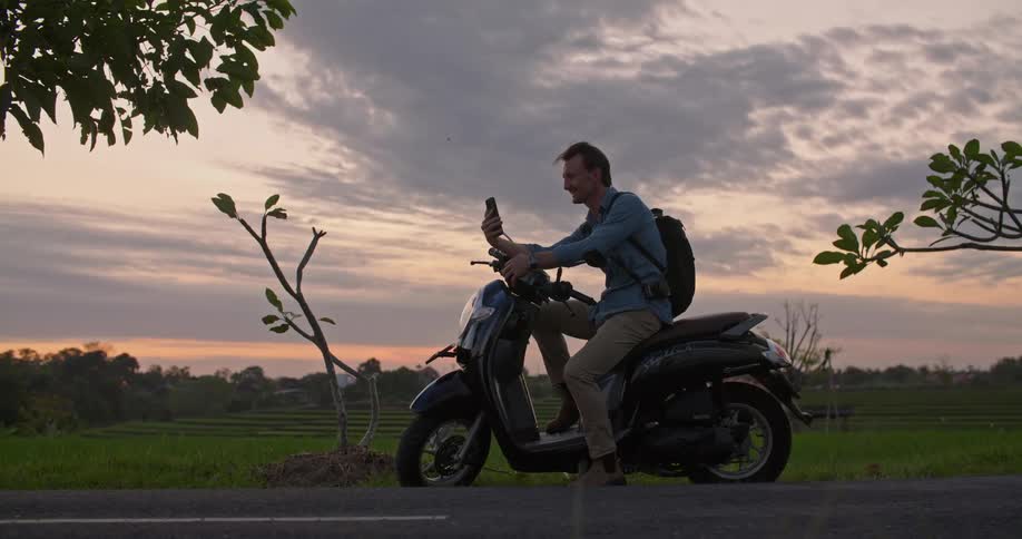 Man with blue shirt brown pants and black backpack sits on moped talks on smartphone in the countryside