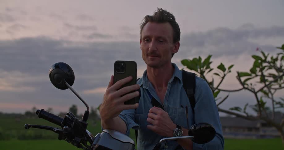 Man sitting on a parked motorcycle on a rural field talking on the phone at evening