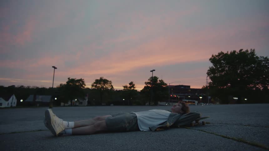 A teenage boy is lying on the pavement of a parking lot with his head on his skateboard