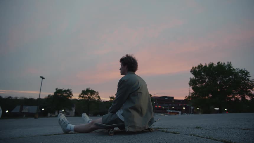 Male skateboarder sits on skateboard in empty parking lot at dusk